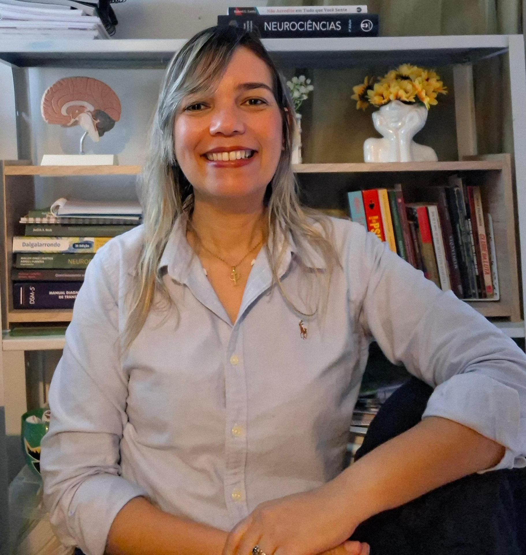 Karla Andréa Souza sorrindo em seu consultório, com estante de livros ao fundo